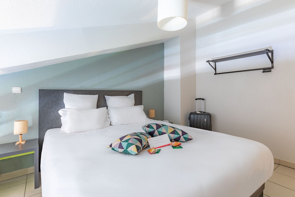 Una cama bien hecha con sábanas blancas y almohadas geométricas de colores, una lámpara de noche y una maleta en el suelo.
