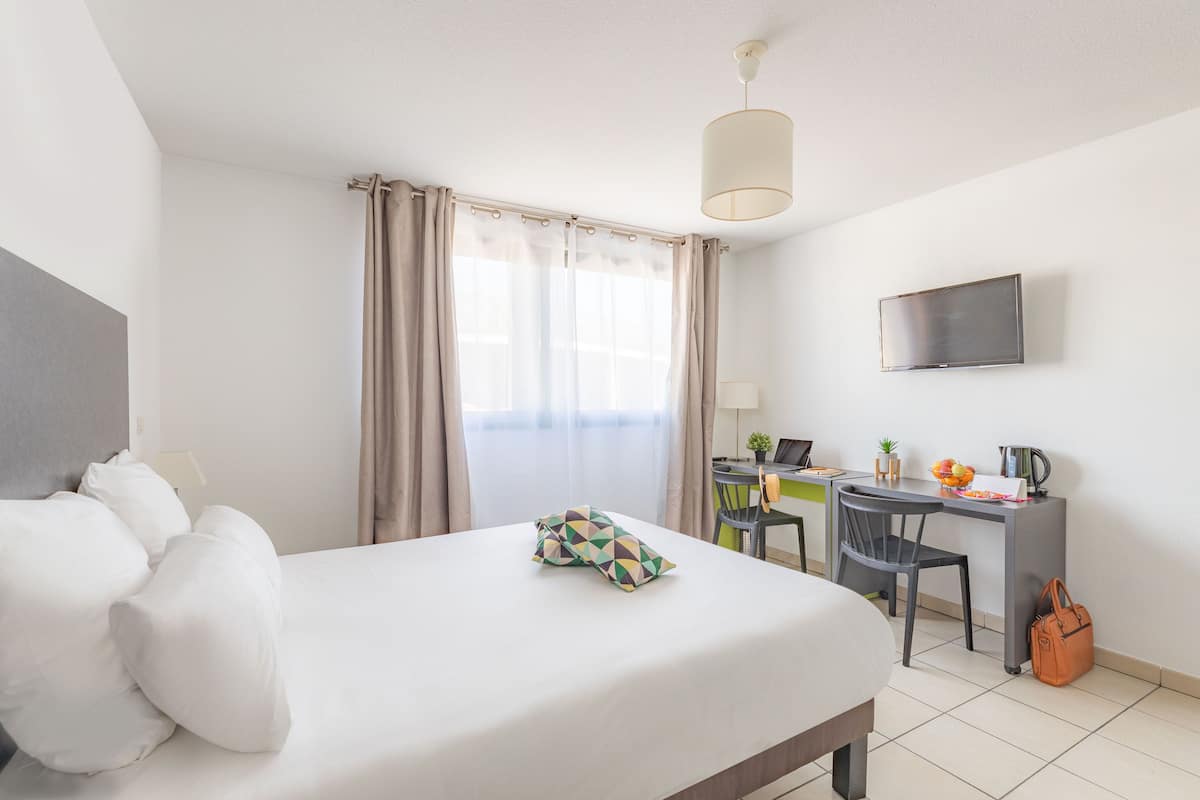 Una habitación de hotel con cama, escritorio, silla, televisor y ventana con cortinas.