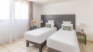 A hotel room with two single beds, a dark headboard, bedside tables with lamps, a small plant, and sheer curtains. - Appart'City Confort La Ciotat Cote Port (La Ciotat)