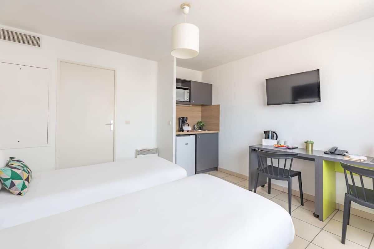 Una habitación de hotel moderna con una cama, un escritorio con una silla, un televisor montado en la pared y una kitchenette al fondo.