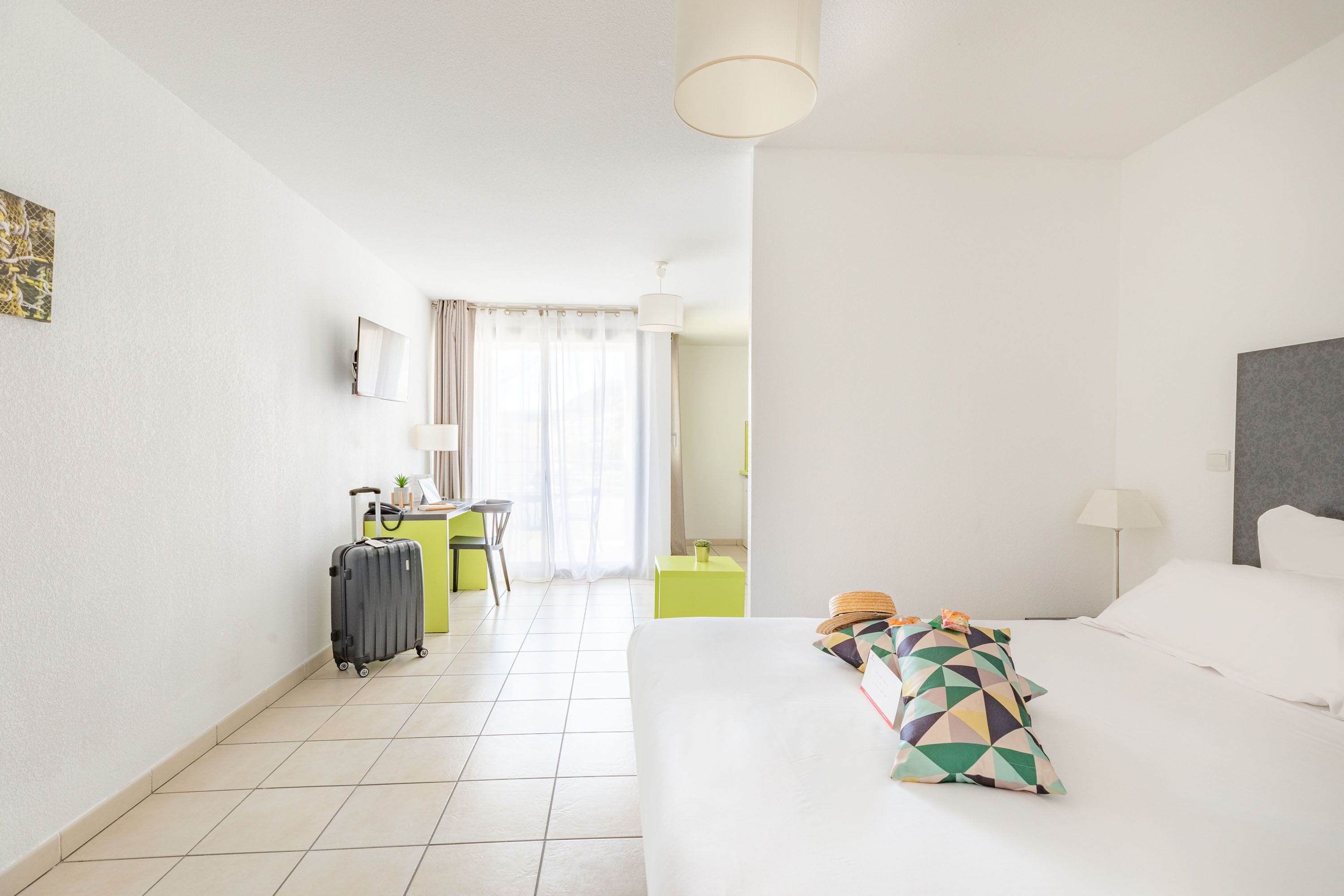 A hotel room with a bed, a desk, a chair, a suitcase, and a small table with a plant.