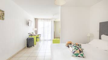 A hotel room with a bed, a desk, a chair, a suitcase, and a small table with a plant.