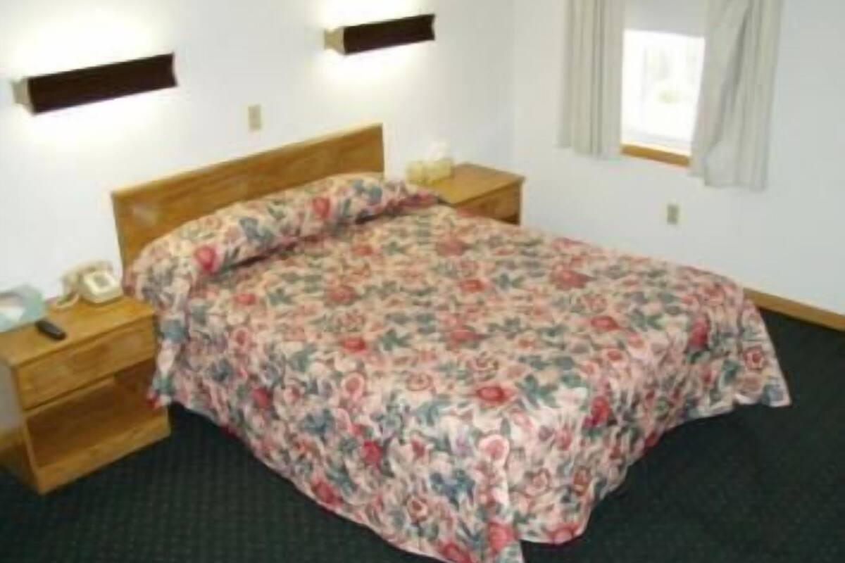 A bedroom with a wooden headboard, a floral bedspread, a bedside table with a lamp, and a window with curtains.