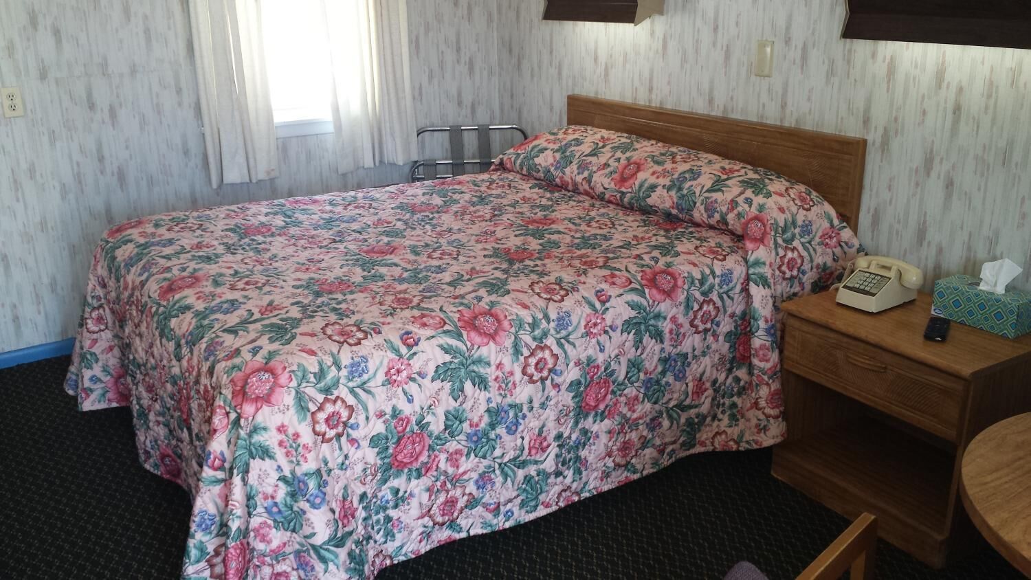 Habitació d'hotel amb llençols estampats florals, una tauleta de nit de fusta amb un telèfon i una taula rodona petita.