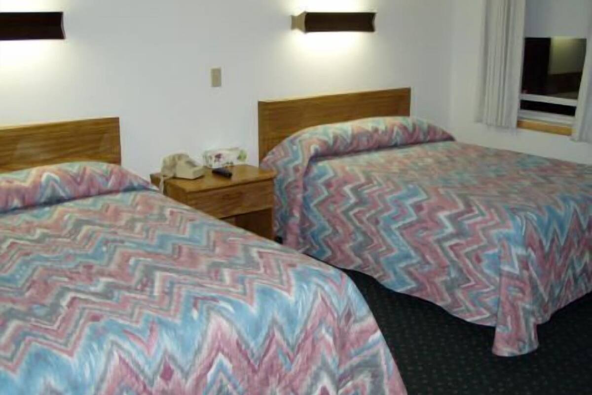 A hotel room with two beds, each with a patterned bedspread, a wooden headboard, a bedside table with a lamp, and a window with curtains.