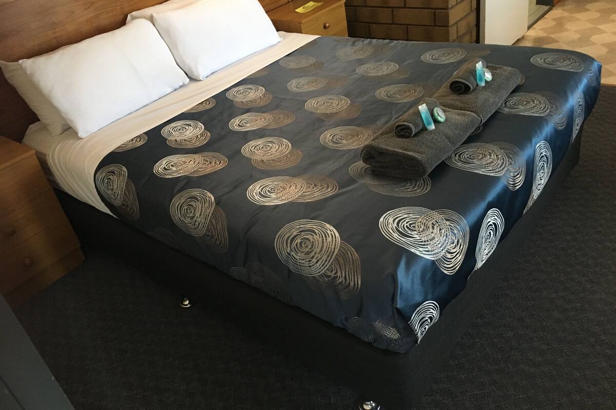 A neatly made bed with a patterned bedspread, white pillows, and a rolled-up towel with a blue and white striped towel on top.