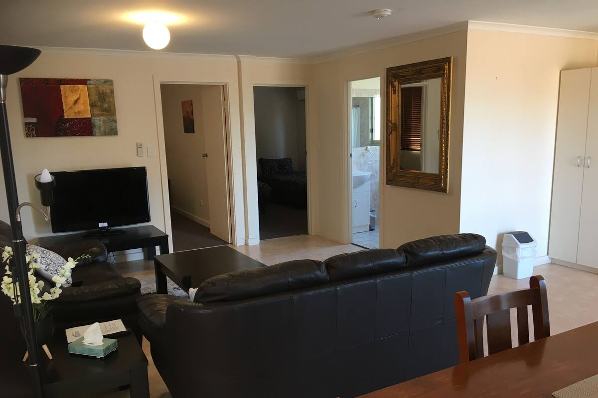 A living room with a black leather sofa, a TV, and a dining table with chairs. There is a hallway leading to a bathroom and a kitchen.