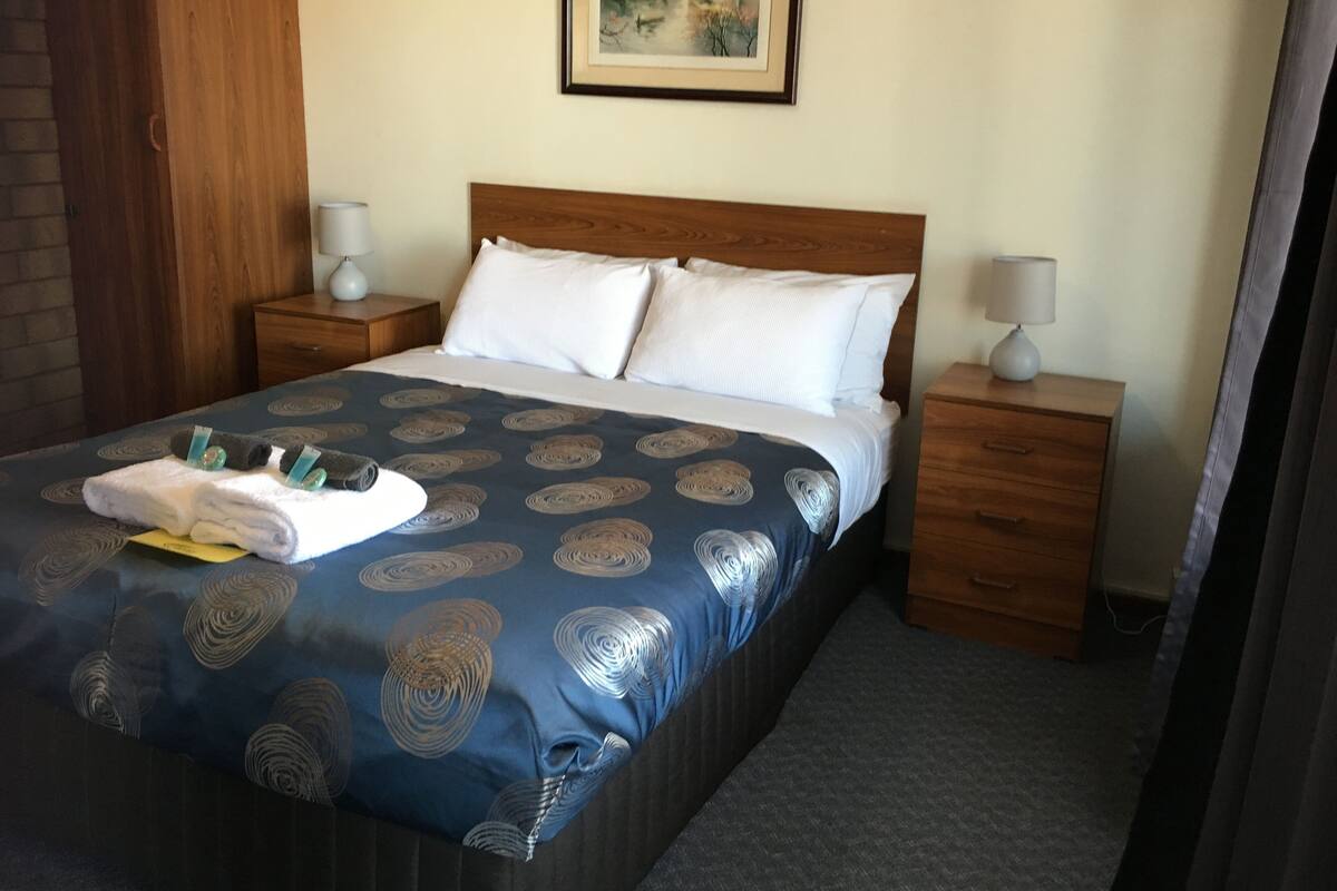A neatly made bed with a blue patterned bedspread, two white pillows, and a folded towel with a yellow note. There are two wooden nightstands with lamps, a wooden wardrobe, and a framed picture on the wall.