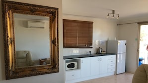 A compact kitchen with white cabinets, a built-in oven, and a refrigerator. A large ornate mirror hangs on the wall, and a beige armchair is in the foreground. - Airport Whyalla Motel (Whyalla Norrie)