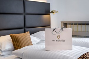 Quarto de hotel com cama, almofadas, cabeceira e uma bolsa branca com texto.