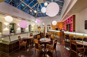 Um lobby de hotel moderno com teto de vidro, mesas e cadeiras redondas, área de bar e escadaria.
