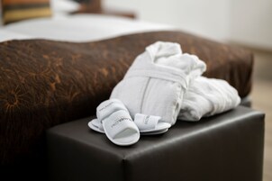 Cama com cobertura marrom estampada, roupão branco e chinelos sobre um banquinho preto.