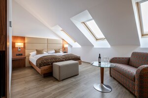 Quarto moderno com teto estilo loft, cama com cabeceira, sofá, mesa de vidro e um pequeno banquinho.