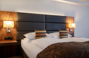 Quarto de hotel moderno com uma cama grande, duas luminárias de cabeceira e um painel de cama de madeira.