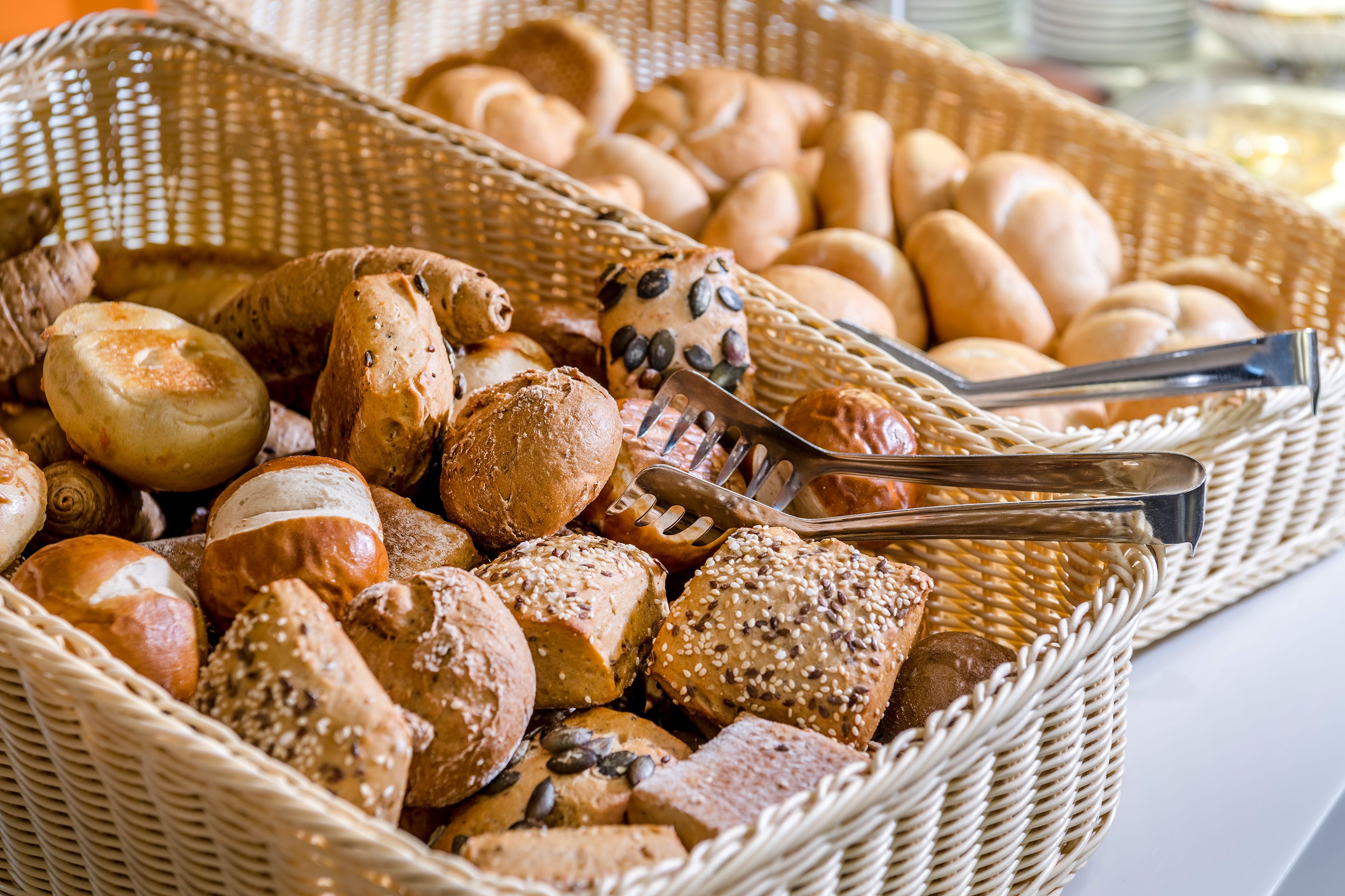 A wicker basket filled with various types of bread, including rolls and loaves, with a pair of tongs resting on top.