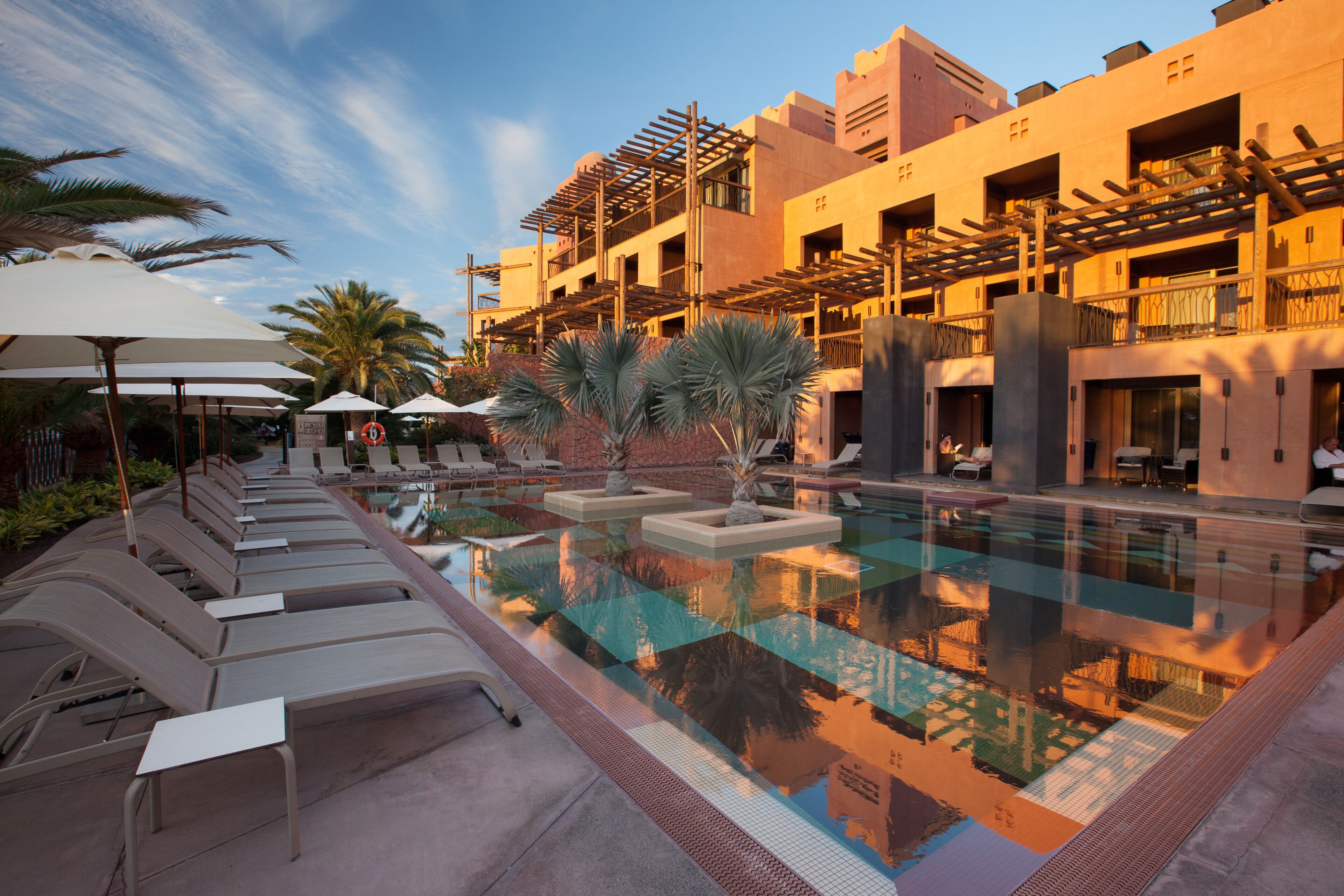A poolside area with lounge chairs and umbrellas, a clear pool reflecting the surroundings, and a multi-story building with balconies.