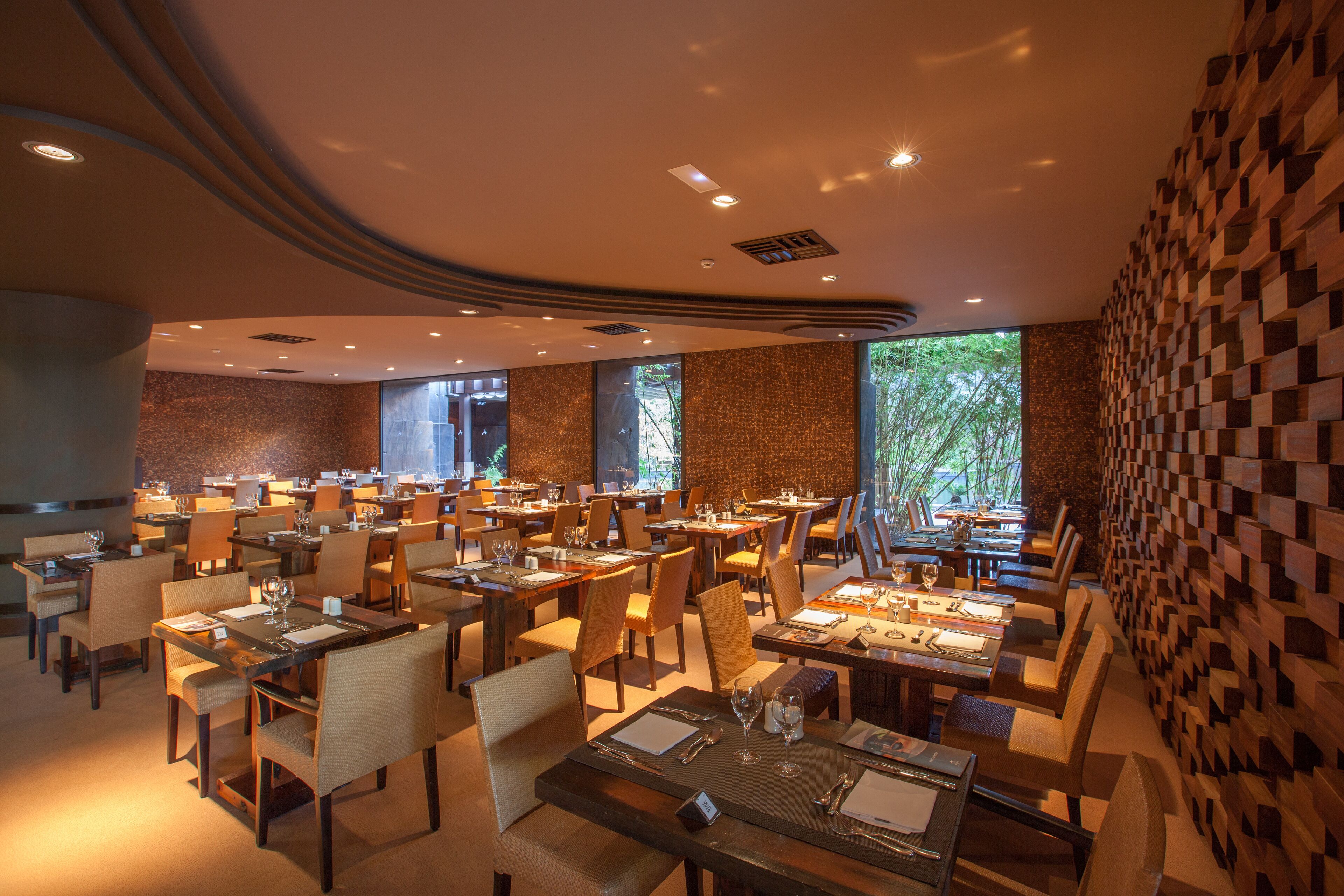 A spacious restaurant with multiple tables and chairs, large windows, and a modern ceiling design.