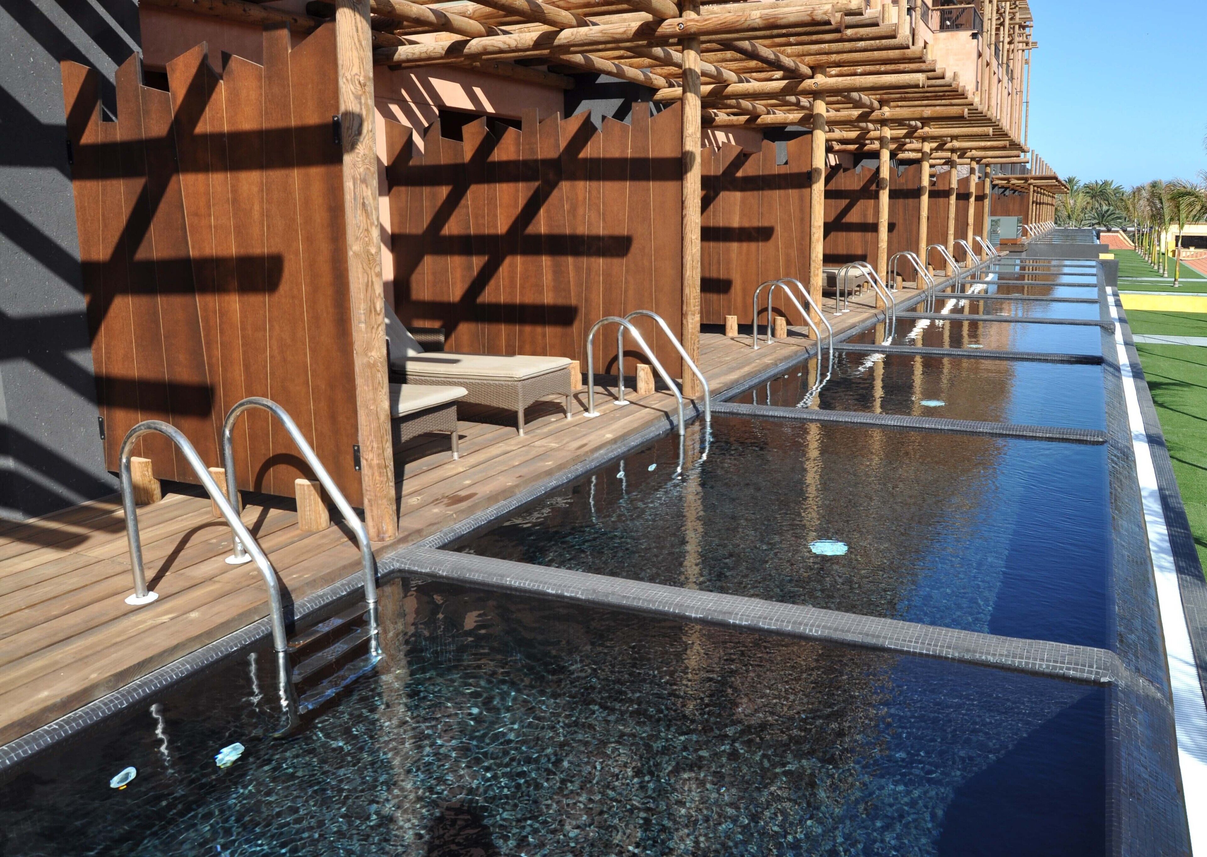 A long, narrow swimming pool with wooden deck seating and metal handrails.