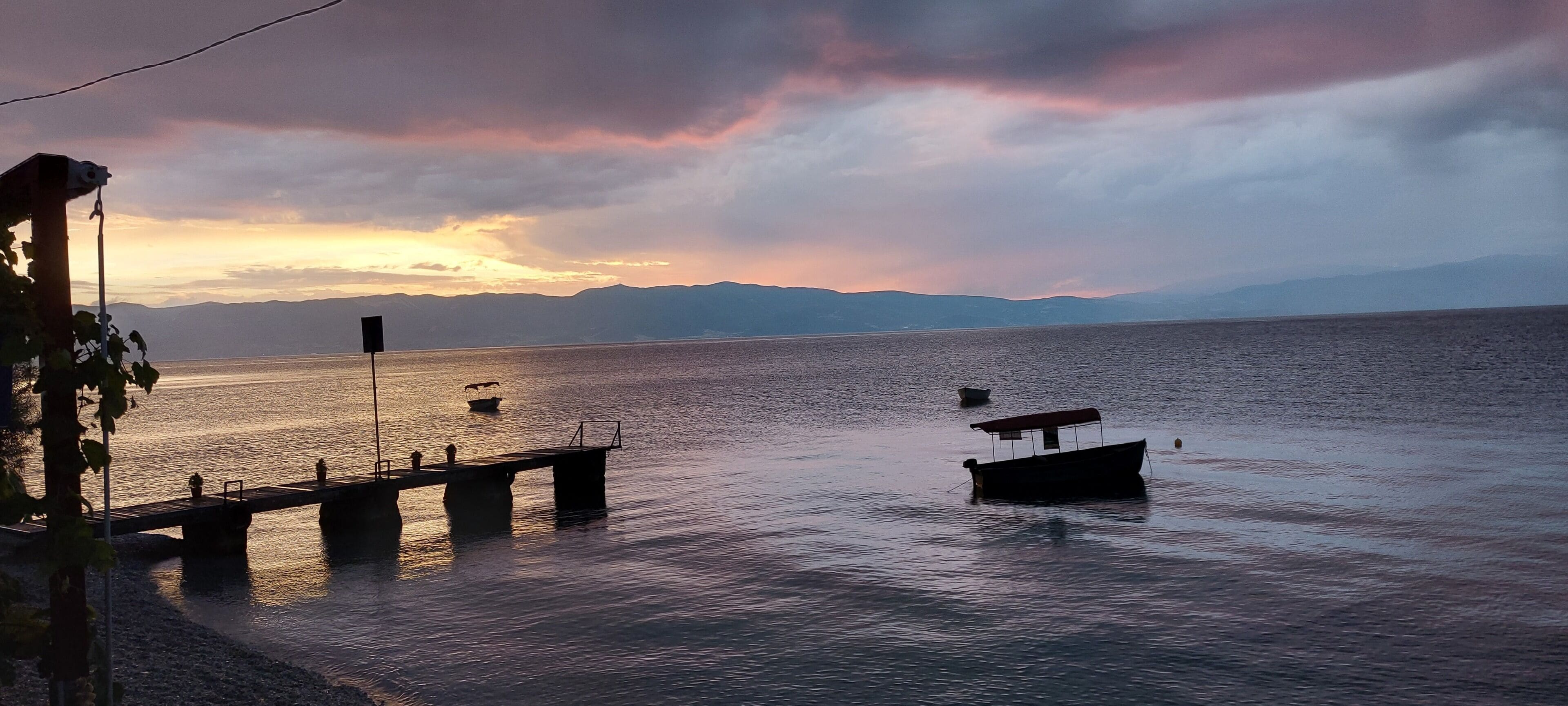 Un lago tranquilo con barcos, un muelle y montañas a lo lejos.