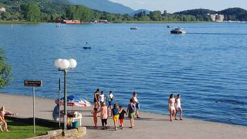 Un paseo marítimo junto al lago con gente caminando y sentada, un cartel con indicaciones y barcos en el agua.