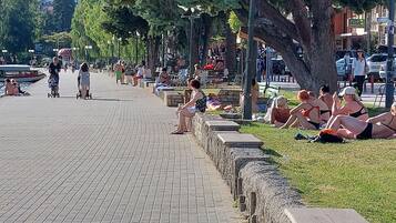 Une promenade le long de la rivière où l’on peut voir des gens se détendre, un bateau amarré sur l’eau et une zone herbeuse avec des bancs.