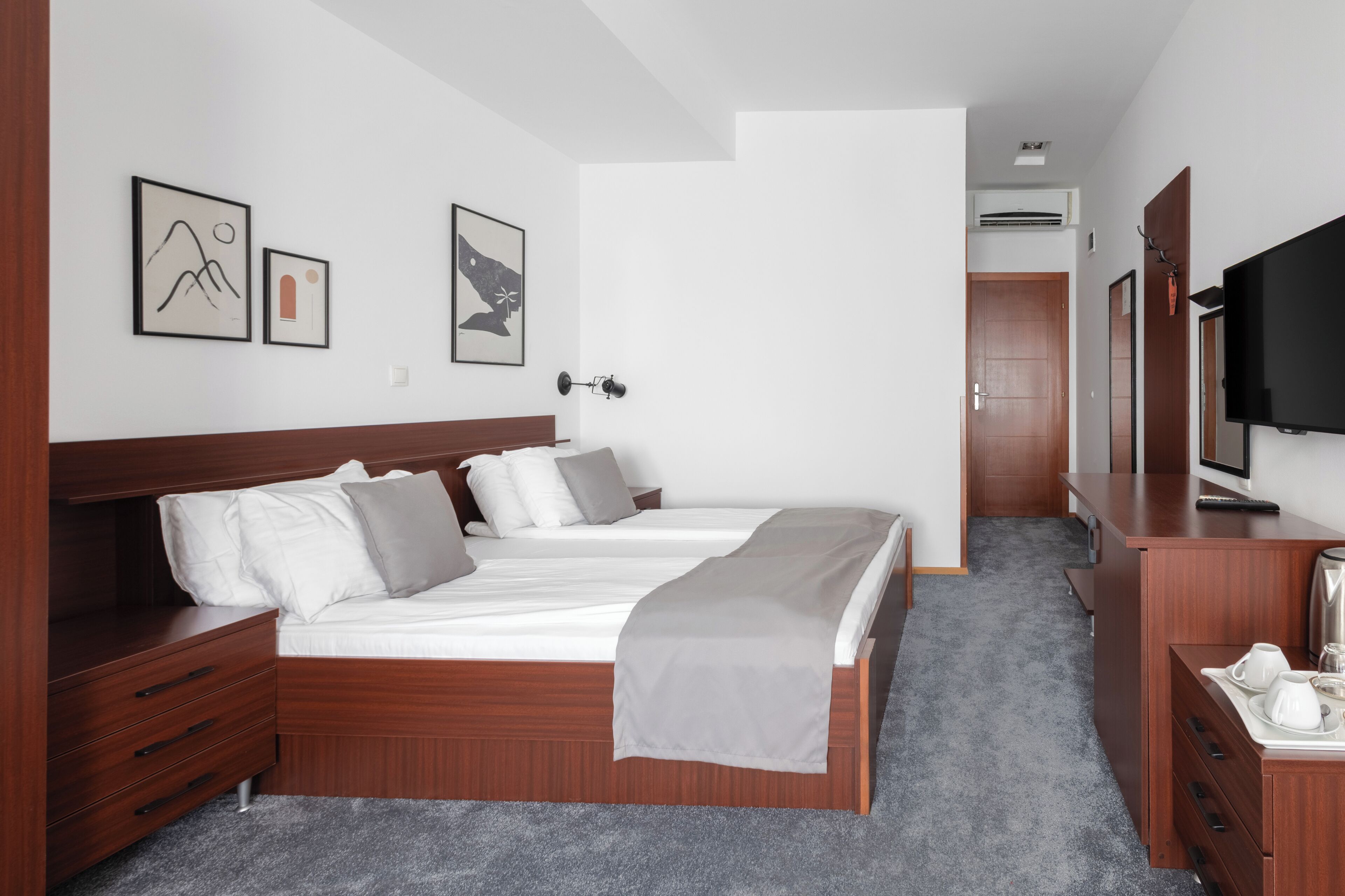 Quarto de hotel com cama de casal, cabeceira de madeira, mesas de cabeceira, televisão e obras de arte nas paredes.