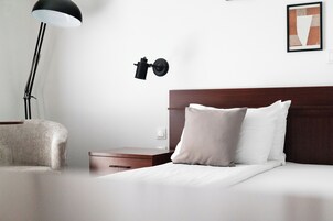 Una cama bien hecha con sábanas blancas y dos almohadas, una mesita de noche con detalles de madera, una lámpara de pie negra y un aplique de pared.