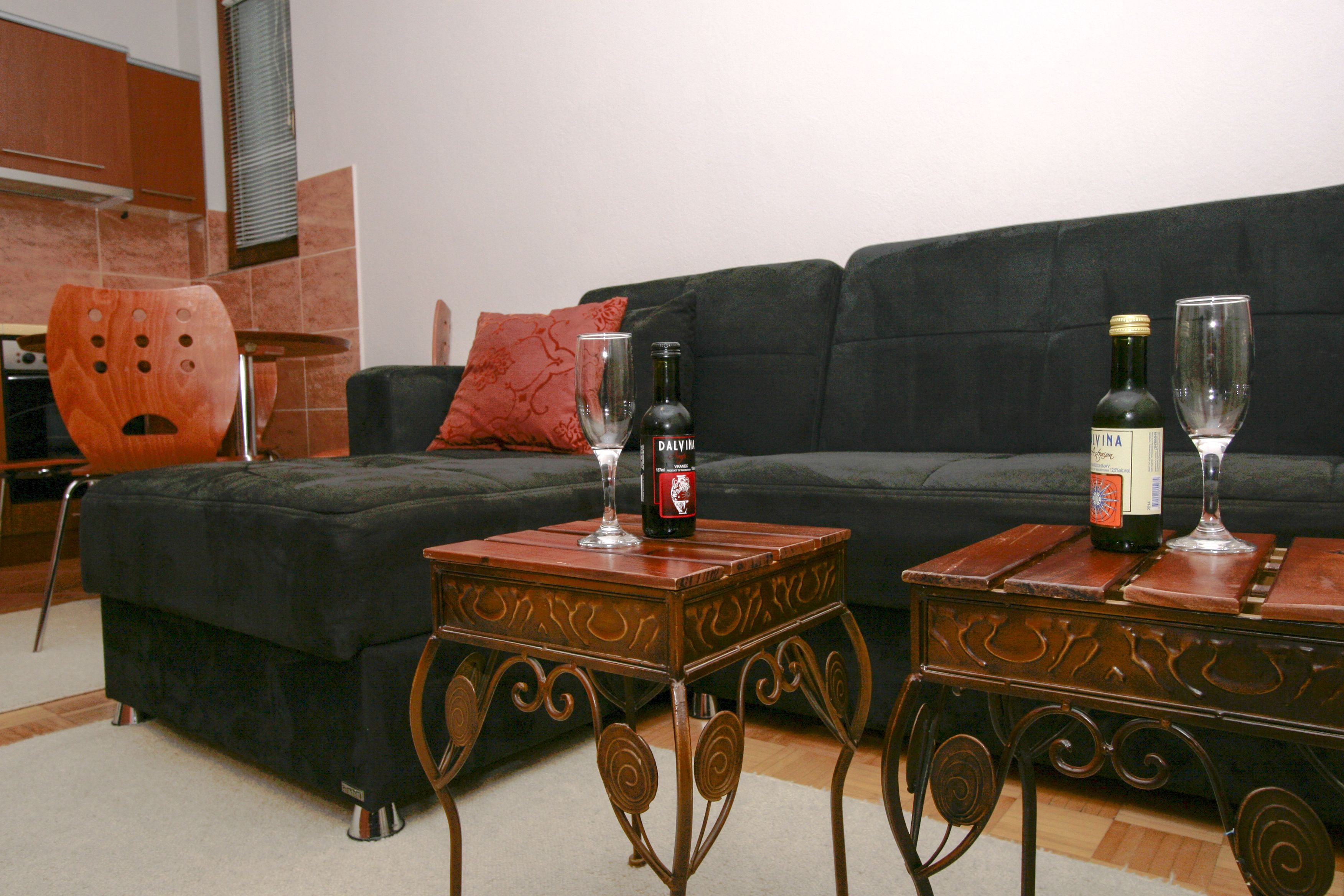 A living room with a dark green sofa, two wooden side tables with wine glasses and bottles, a red patterned pillow, and a wooden chair.
