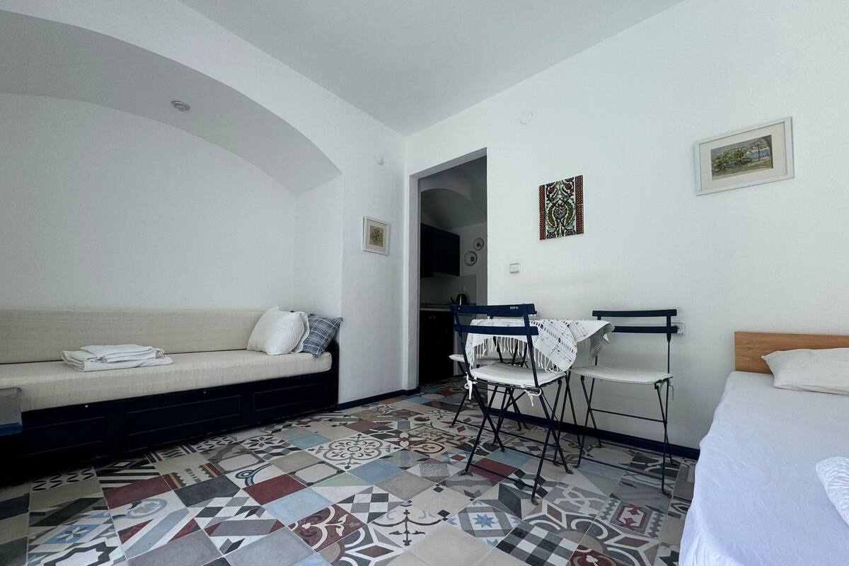 Una habitación con un sofá, una pequeña mesa con sillas y una cama. El piso tiene un diseño de baldosas estampado.