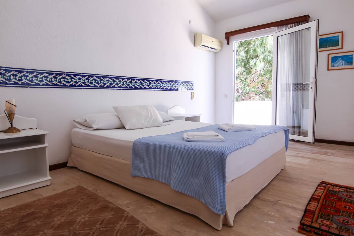 Un dormitorio con cama, mesita de noche, lámpara y ventana con vistas a zonas verdes.