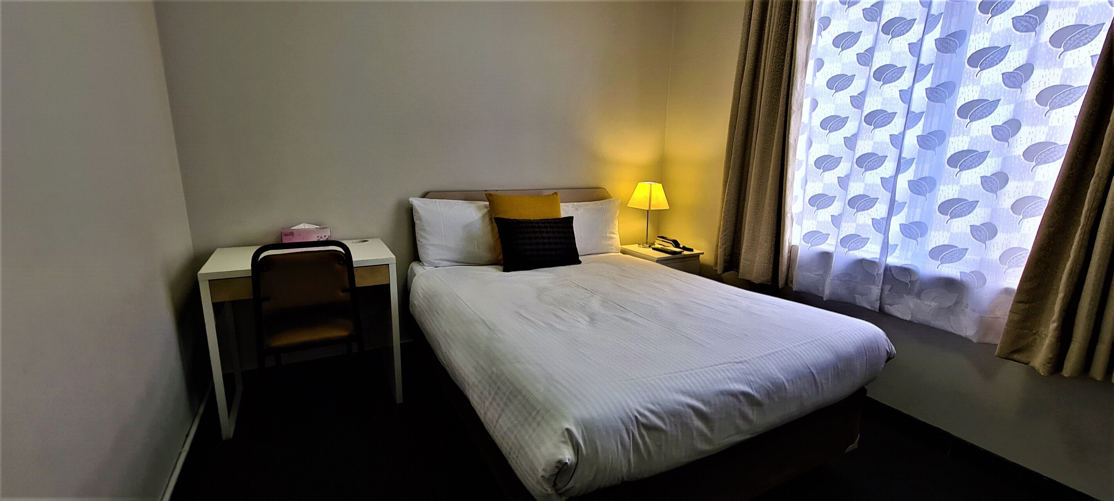 A hotel room with a bed, a desk, a chair, a lamp, and a window with curtains.