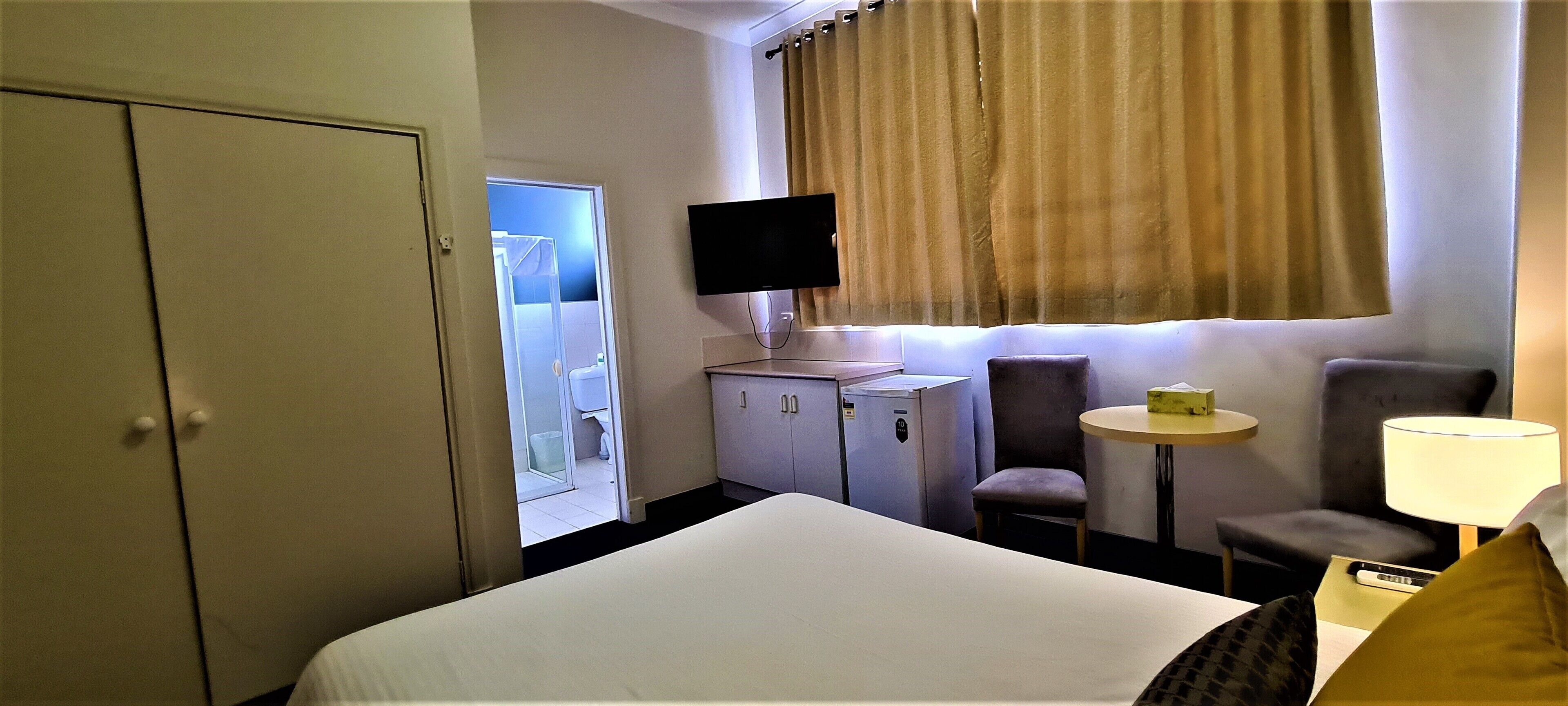 Habitación de hotel con una cama, un escritorio con computadora, una silla, un televisor montado en la pared y una ventana con cortinas.