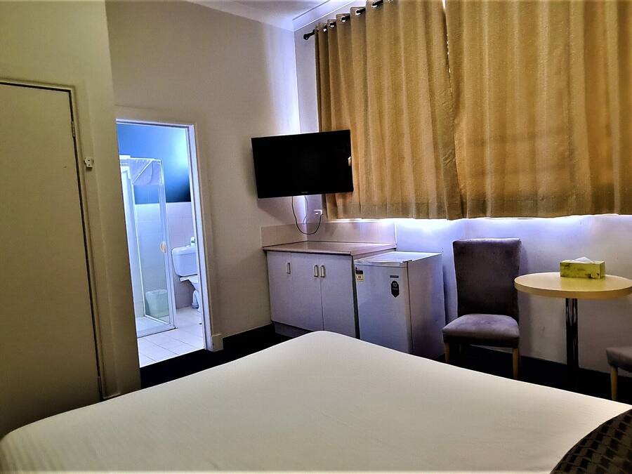 Habitación de hotel con una cama, un escritorio con computadora, una silla, un televisor montado en la pared y una ventana con cortinas.