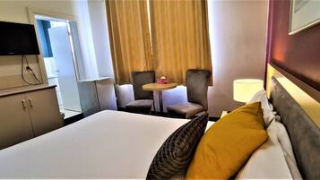 Habitación de hotel con cama, dos sillones, una mesita con lámpara, televisor en la pared y ventana con cortinas.