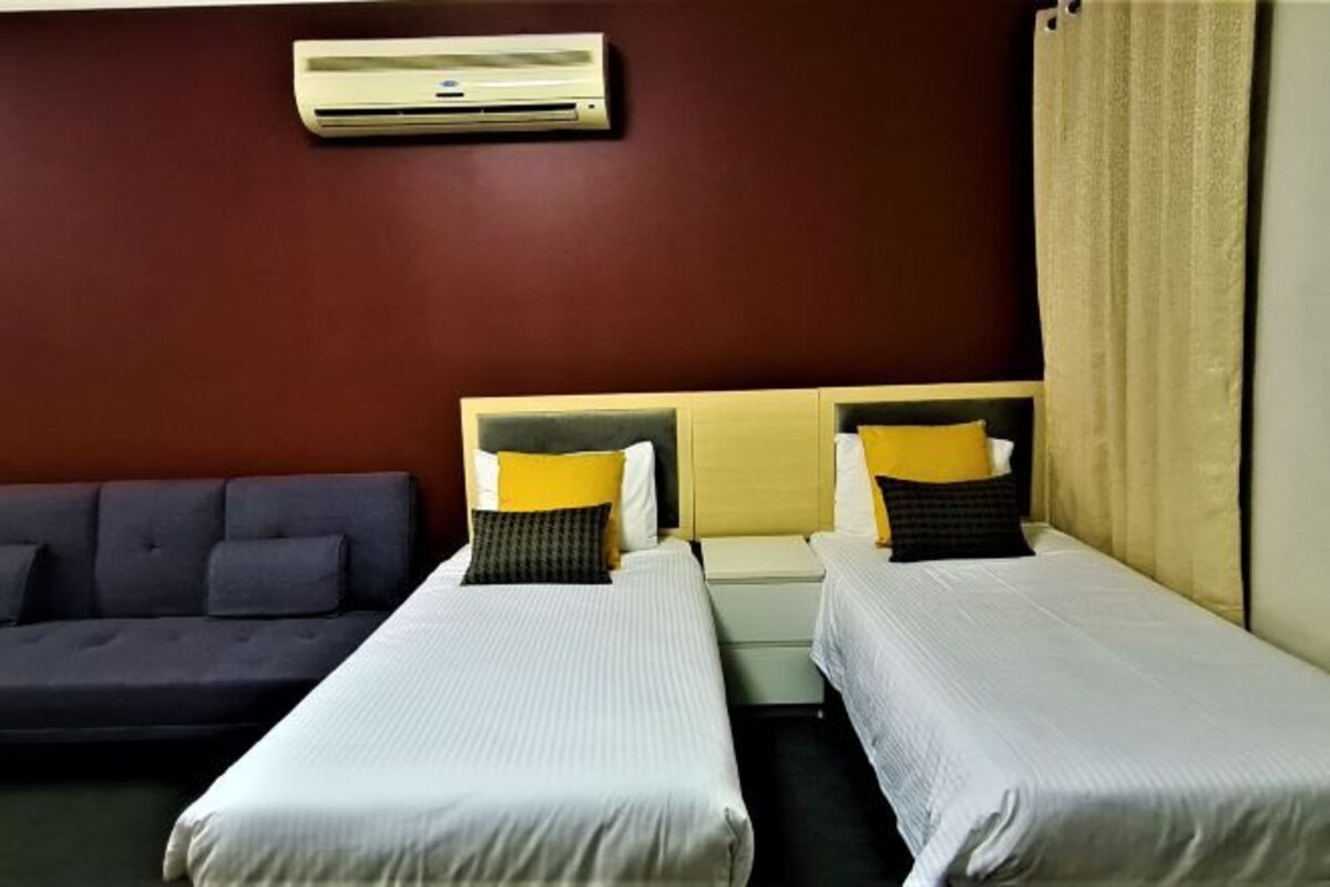 A hotel room with two single beds, a sofa, an air conditioning unit, and a curtain.