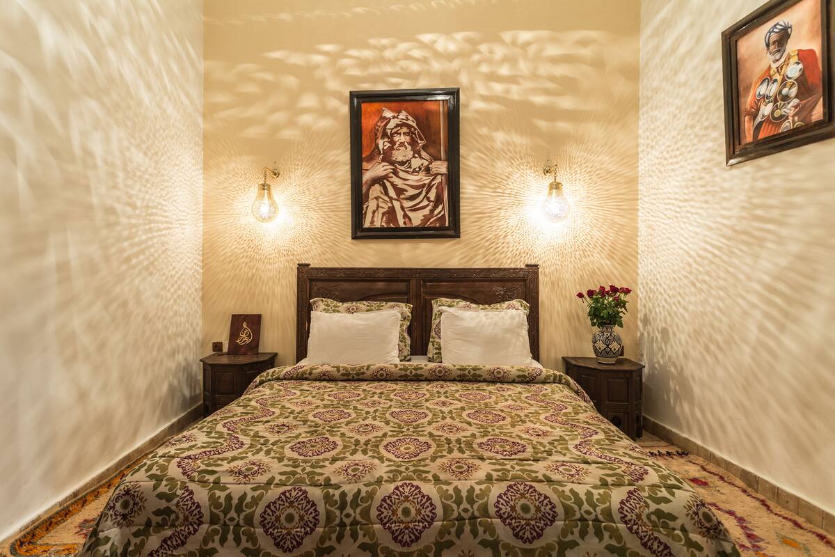 Cama bem arrumada com colcha estampada, duas mesas de cabeceira com abajures, uma obra de arte emoldurada acima da cama e um vaso com flores em uma das mesas.