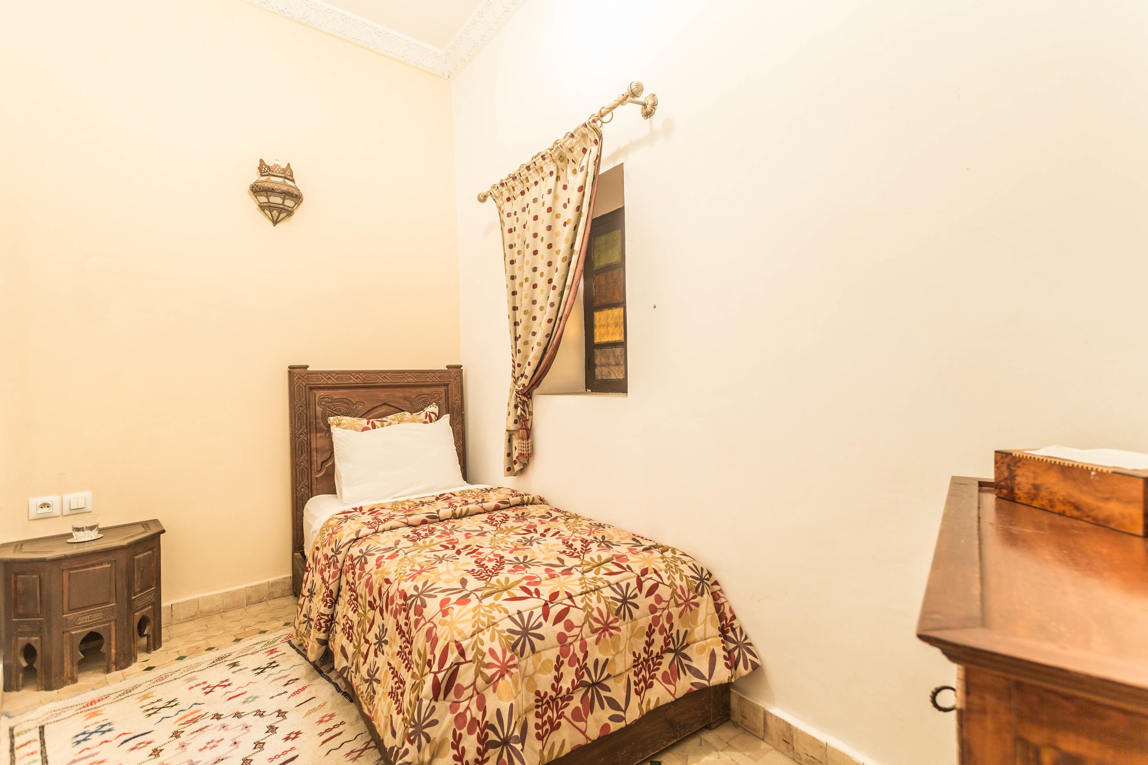 Un dormitorio con cama, mesita de noche, cómoda de madera, alfombra estampada y lámpara fijada a la pared.