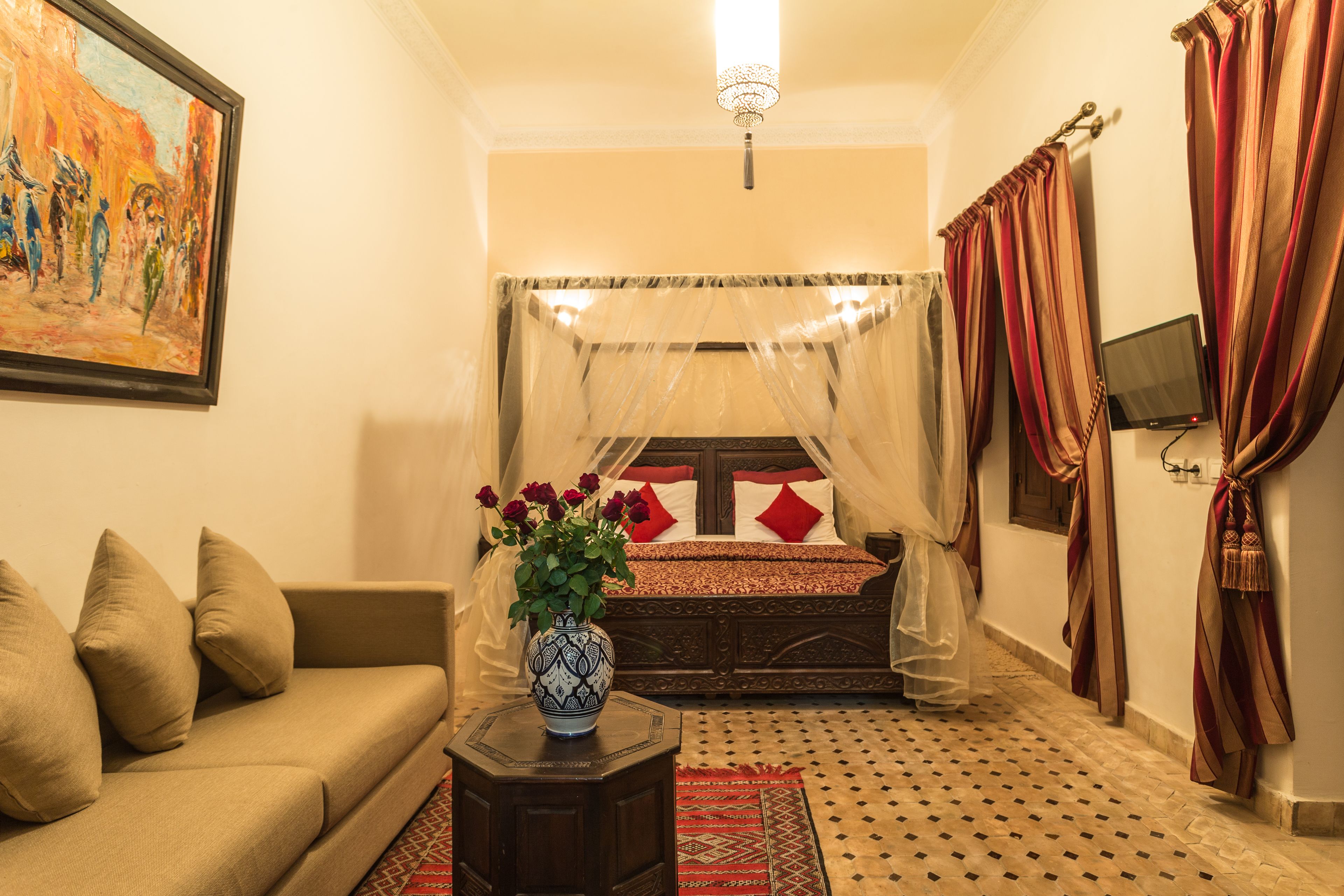 A room with a bed, a sofa, a vase with flowers, a painting, and a television.
