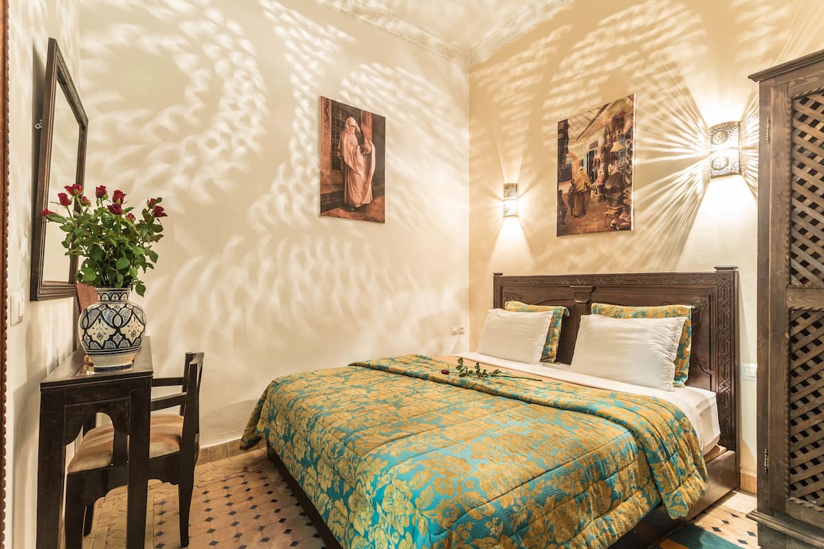 Quarto com cama, mesa de cabeceira, cadeira, vaso com flores e duas pinturas penduradas na parede.