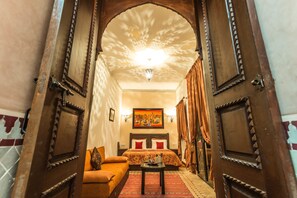 A hotel room with a wooden door, a bed with a floral bedspread, a sofa, and a small table with a lamp.