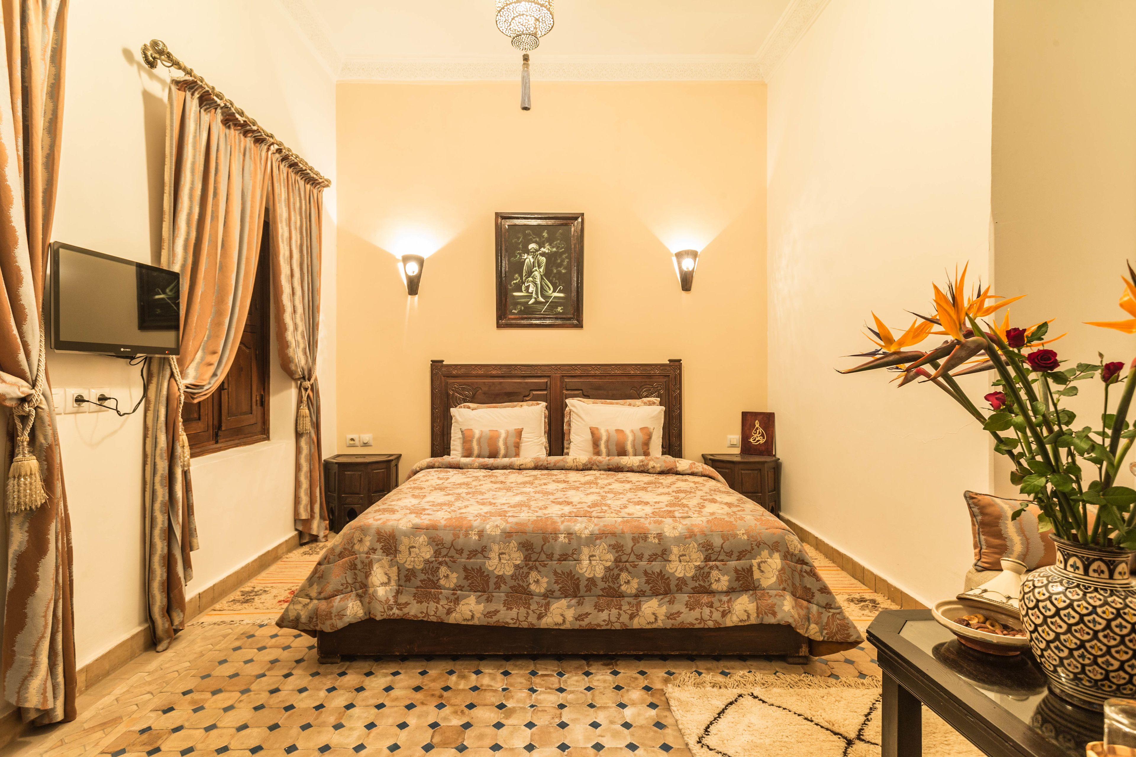 Un dormitorio con una cama, dos lámparas de pared, un televisor de pantalla plana, un jarrón decorativo y una alfombra estampada.