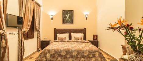 Un dormitorio con una cama, dos lámparas de pared, un televisor de pantalla plana, un jarrón decorativo y una alfombra estampada.