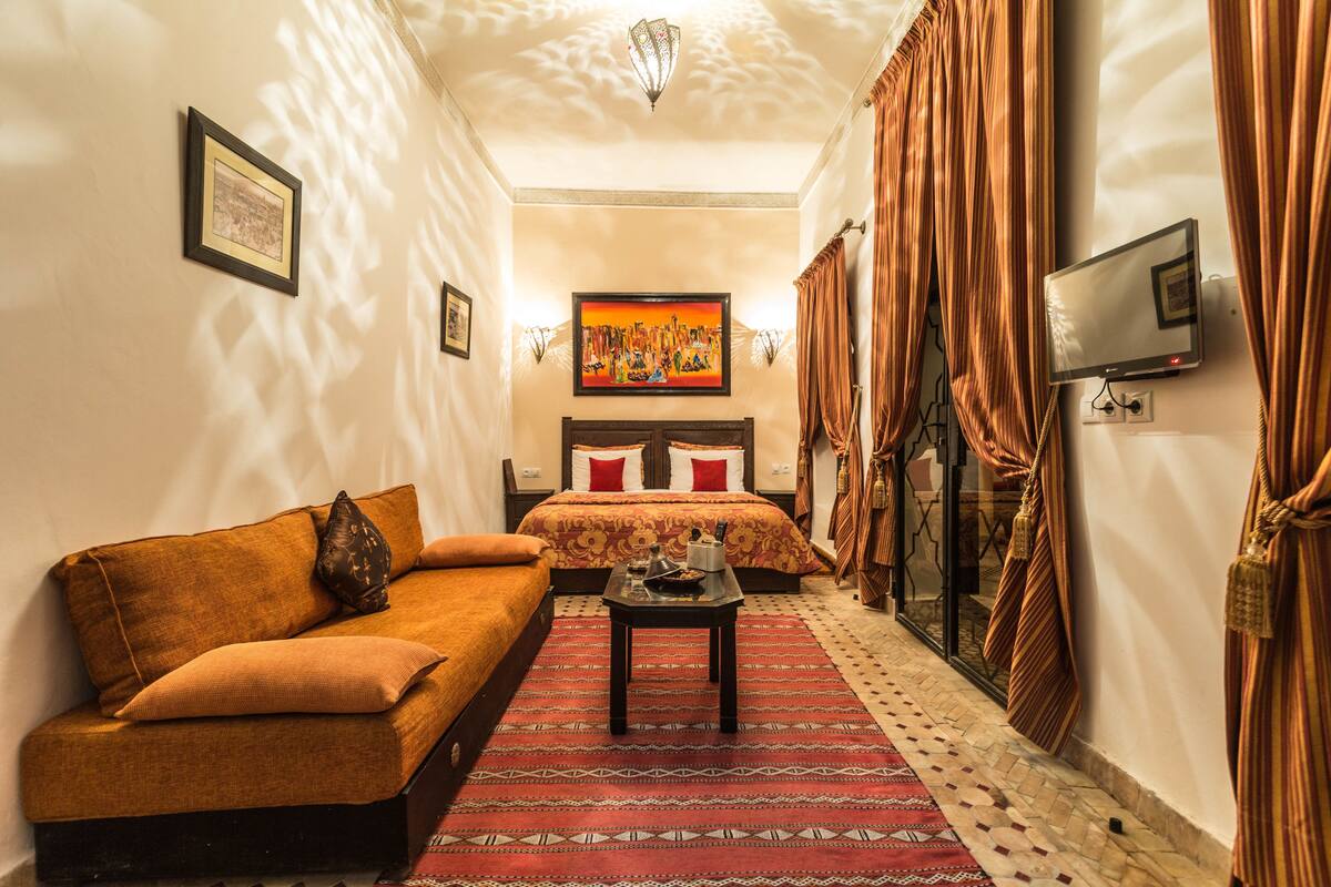 Quarto de hotel com sofá, cama com colcha estampada, mesa pequena com abajur e televisão fixada na parede.
