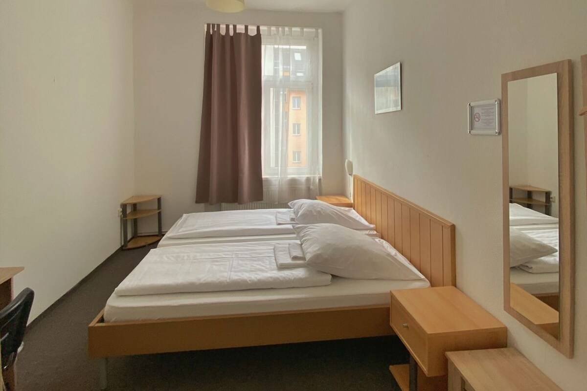A hotel room with a bed, bedside tables, a mirror, a window with curtains, and a hanging light fixture.