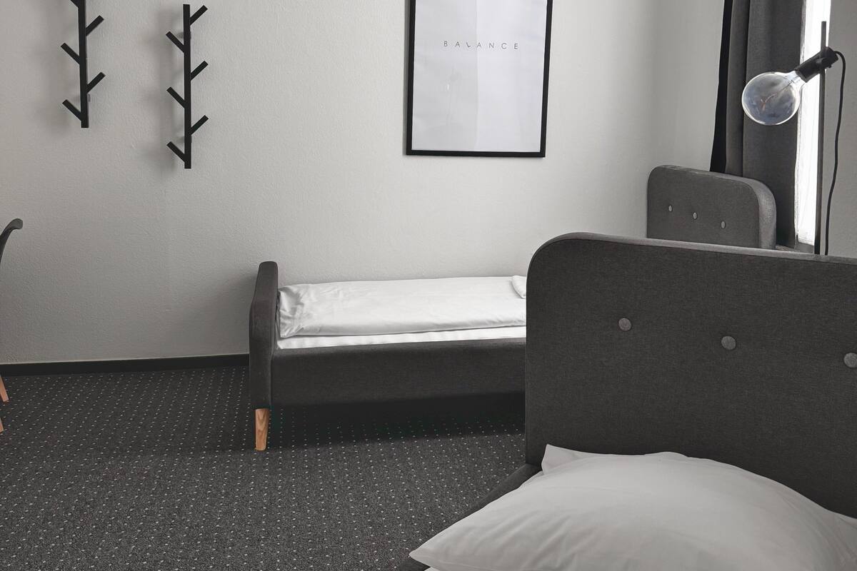 A minimalist hotel room with a single bed, a framed artwork on the wall, and a unique ceiling light fixture.