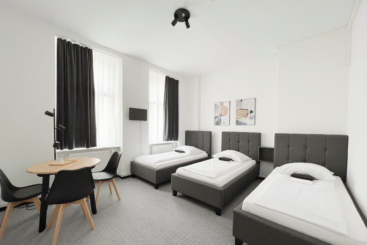 A hotel room with two single beds, a sofa bed, a desk with a chair, and a TV on the wall.