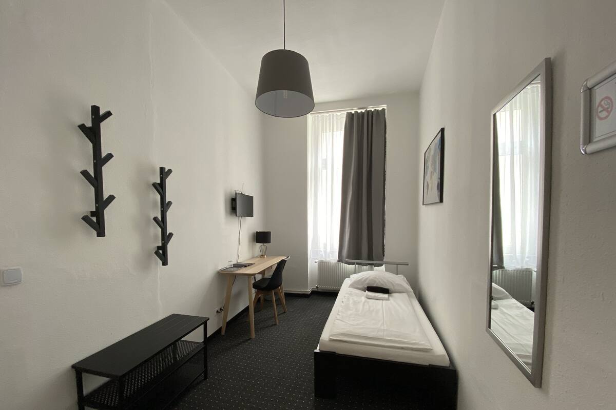 A compact hotel room with a single bed, a desk with a chair, a small table, a TV, and a large mirror.