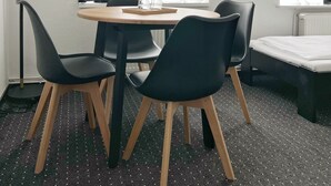 A modern room with a round wooden table, black chairs, a floor lamp, and a bed. - Hotel AVAlex (Berlin)