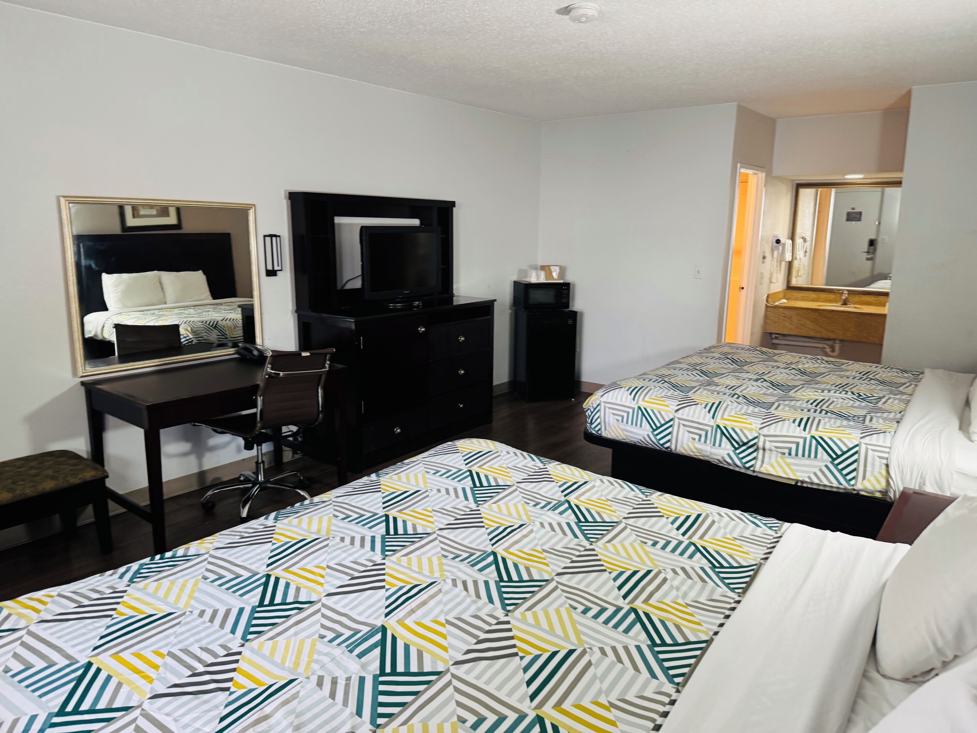 A hotel room with two beds, a desk, a chair, a dresser, a TV, and a bathroom visible through an open door.