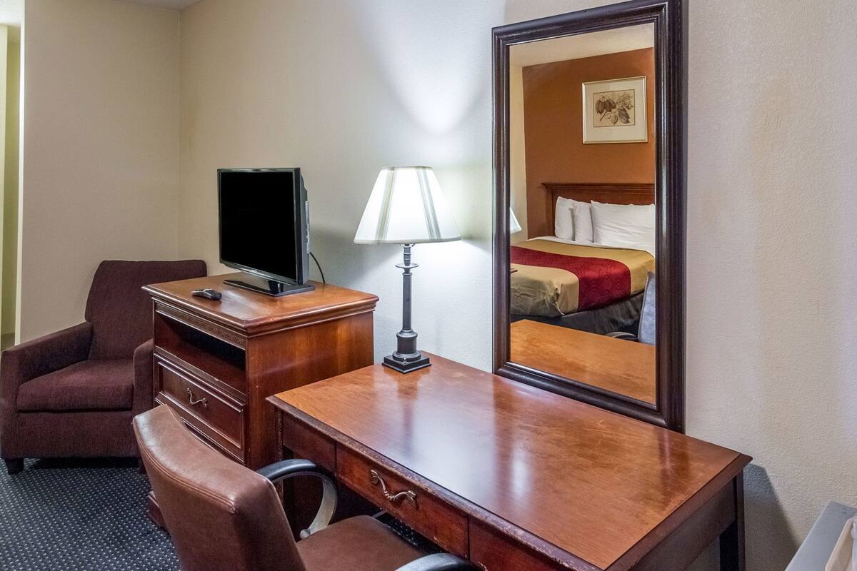 A hotel room with a wooden desk, a television, a lamp, a chair, and a bed with a red and beige bedspread.